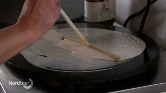 Crêpe-Teig wird mit einem Holzstäbchen dünn auf einer Herdplatte verstrichen. © Screenshot 
