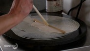 Crêpe-Teig wird mit einem Holzstäbchen dünn auf einer Herdplatte verstrichen. © Screenshot 