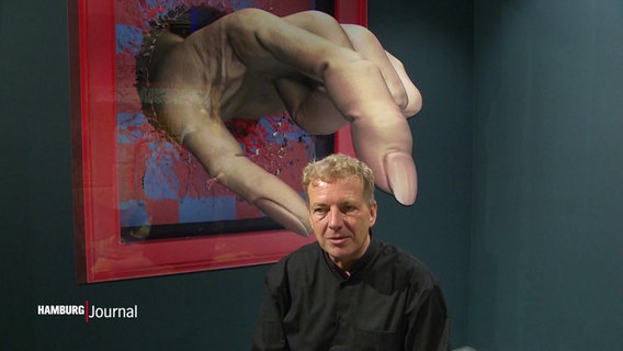 Ein Mann sitzt auf einem Stuhl während sein Kopf scheinbar von einer überdimensionalen Hand, die aus der Wand ragt, erfasst wird. © Screenshot 