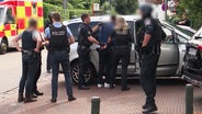 Polizeikräfte in schusssicheren Westen führen eine Person in Handschellen zu einem Fahrzeug. © Screenshot 