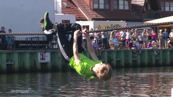 Ein junger Mann hangelt sich mit Armen und Beinen ein Seil entlang. © Screenshot 