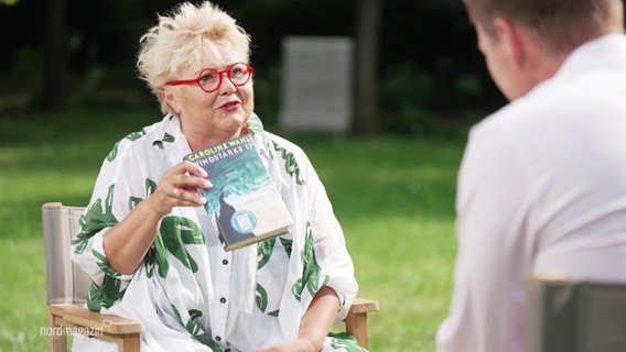Eine Frau sitzt in einem Garten und hält ein Buch in den Händen. © Screenshot 