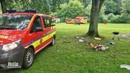 In einem Park steht ein Sanitäter-Notfallfahrzeug. Daneben zurückgelassene Dinge für einen Parkbesuch: Darunter ein Kinderfahrrad und ein Grill. © Screenshot 