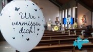 In einer Kirche stehen Blumen und Fotos zum dedenken an einen toten Jungen, im Vordergrund sieht man einen Ballon mit der Aufschrift: "Wir vermissen dich!". © Screenshot 