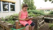 Ein Mädchen sitzt auf der Kante eines Hochbeetes und liest aus einem Buch vor. © Screenshot 
