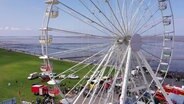 Ein großes weißes Riesenrad ragt über das Festgelände der Feuerwehrtage auf. Im Hintergrund ist das Wattenmehr zu sehen. © Screenshot 