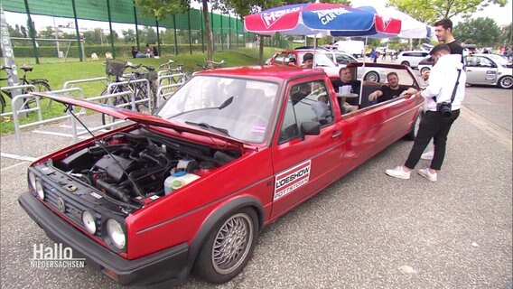 Fans mit Getränken sitzten in einem umgebauten GTI-Modell mit geöffneter Motorhaube  unter einem Sonnenschirm. © Screenshot 
