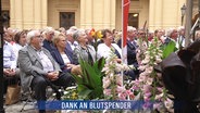 Festlich gekleidetes Publikum in einem mit Blumen geschmückten Saal. darunter die Schlagzeile: "Dank an Blutspender" © Screenshot 
