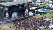 Blick aus der Vogelperspektive auf eine Menschenmenge vor einer größeren Open-Air-Bühne auf einem Hard-Rock-Festival. © Screenshot 