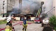 Feuerwehrleute löschen eine brennende Lagerhalle zwischen Getreidesilos. © Screenshot 