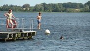 Ein kleiner Junge hüpft auf dem Sprungbrett einer schwimmenden Plattform auf einem Badesee auf und ab. © Screenshot 