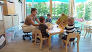 Eine Erzieherin sitzt mit 3 spielenden Kindern an einem Tisch. © Screenshot 