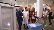 Zwei Männer und eine Frau stehen um den Arm eines Operationsroboters herum. © Screenshot 