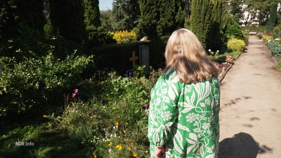 Eine Frau steht vor einer Grabstelle, auf der Gräser und Wildblumen wachsen. © Screenshot 