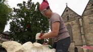 Eine Bildhauerin bearbeitet auf einem Kirchplatz ein großes Stück Holz. © Screenshot 