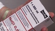 Eine Person hält eine Schachtel mit Kochsalzlösung in der Hand. © Screenshot 