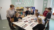 Eine Gruppe Kinder und eine Lehrerin stehen um einen Tisch auf dem Bilder von hypothetischen Schulhofgestaltungen liegen. © Screenshot 