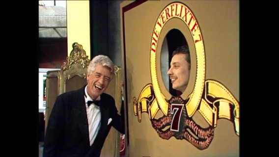 Moderator Rudi Carrell und ein Mann, der durch ein Schild "Die verflixte 7" schaut. © Screenshot 