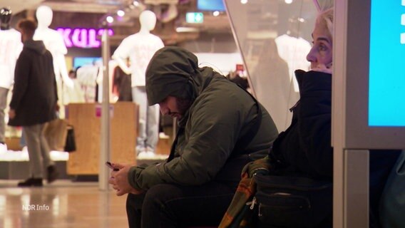 Ein Mann mit übergezogener Kapuzenjacke sitzt mit krummen Rücken in einem Einkaufszentrum sein Smartphone haltend. © Screenshot 