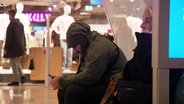Ein Mann mit übergezogener Kapuzenjacke sitzt mit krummen Rücken in einem Einkaufszentrum sein Smartphone haltend. © Screenshot 