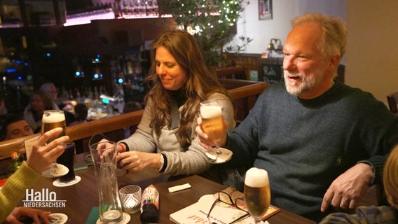 Mehrere Personen sitzen in einem Restaurant, der Mann prostet mit einem Bier seinem Gegenüber zu. © Screenshot 