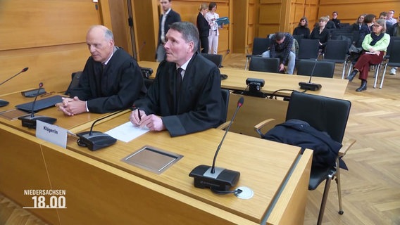 Zwe Männer sitzen in einem Gerichtssaal am Tisch des Klägers, neben ihnen ein freier Stuhl. © Screenshot 