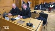 Zwe Männer sitzen in einem Gerichtssaal am Tisch des Klägers, neben ihnen ein freier Stuhl. © Screenshot 