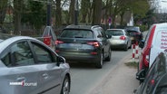 Ein Autoschlange fährt in langsamem Tempo an dem Eingang zu einem Schulgeländer in einer Einbahnstraße entlang. © Screenshot 