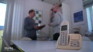 Ein Telefon steht auf einem Tisch - im Hintergrund diskutieren zwei Männer. © Screenshot 