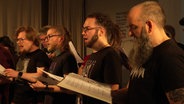 Die Mitglieder eines Metal-Chors stehen mit Notenblättern in der Hand nebeneinander und singen. © Screenshot 