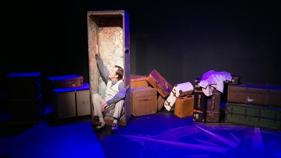 Ein Schauspieler sitzt bei einer Szene eines Theaterstücks in einem größeren Koffer, daneben: weiteres Reisegepäck. © Screenshot 