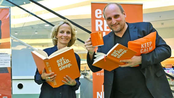 eine Frau und ein Mann stehen nebeeinander, halten orangefarbene Kissen, Bücher und Tassen mit der Aufschrift EAT READ SLEEP und lächeln (NDR Kultur Podcastshosts Katharina Mahrenholtz und Daniel Kaiser) © NDR / Angelika Helm Foto: Angelika Helm