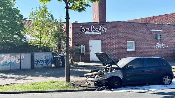 Vor einem Club steht ein ausgebrannter Pkw auf einer Straße. © Screenshot 