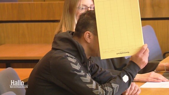 Eine Person hält sich im Gerichtssaal einen Aktenordner vors Gesicht. © Screenshot 
