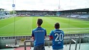 Zwei Personen in Trikots blicken ins Stadion von Holstein-Kiel. © Screenshot 