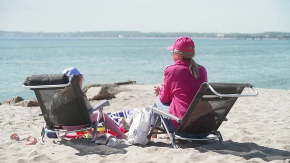 Eine Frau und ein Kind sitzen in Liegestühlen am Strand. © Screenshot 