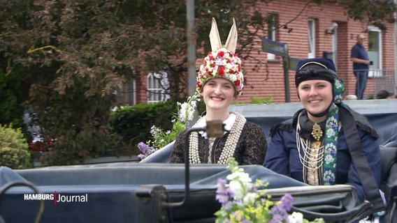 Mit Blumen und Perlenketten geschmückte Frauen sitzen in einer Kursche. © Screenshot 