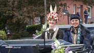 Mit Blumen und Perlenketten geschmückte Frauen sitzen in einer Kursche. © Screenshot 