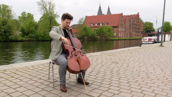 Cellist Gabriel Schwabe sitzt am Traveufer in Lübeck. © Screenshot 