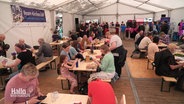 Ein Zelt gefüllt mit vielen Menschen, die an Tischen essen. © Screenshot 