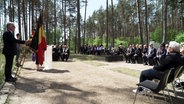 Viele Menschen sitzen im Wald im Kreis vor einem Rednerpult. © Screenshot 