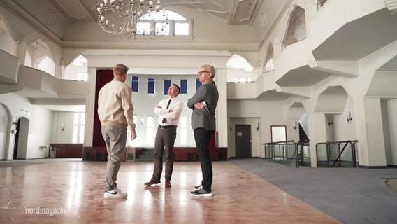 Drei Männer stehen in einem großen Saal mit Kronleuchter. © Screenshot 