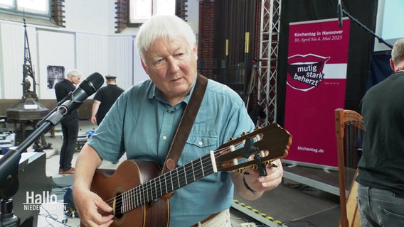 Kirchenliedermacher Fritz Baltruweit stimmt seine Gitarre. Er ist ein Mann mit kurzen grauen Haaren. © Screenshot 