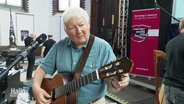 Kirchenliedermacher Fritz Baltruweit stimmt seine Gitarre. Er ist ein Mann mit kurzen grauen Haaren. © Screenshot 