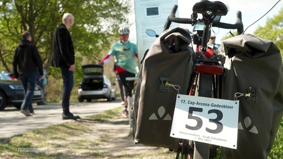 Nahaufnahme von einem Fahrrad, unter dessen Rücklichtern ein Schild angebracht ist. Darauf steht: "17. Cap-Arcona-Gednektour". © Screenshot 