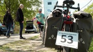 Nahaufnahme von einem Fahrrad, unter dessen Rücklichtern ein Schild angebracht ist. Darauf steht: "17. Cap-Arcona-Gednektour". © Screenshot 