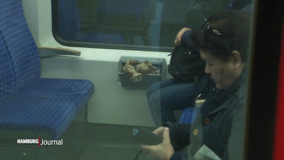 Blick durch die Scheibe einer S-Bahn auf einen offenen und übervollen Müllbehälter zwischen den Sitzen. © Screenshot 