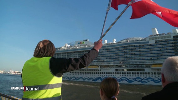 Ein Mann in einer gelben Warnweste schwenkt Flaggen in Richtung eines Kreuzfahrtschiffes auf der Hamburger Elbe. © Screenshot 