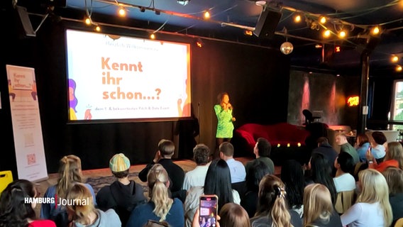 Vor einer Bühne mit Leinwand sitzen zahlreiche Personen und lauschen einer Präsentation. © Screenshot 