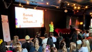 Vor einer Bühne mit Leinwand sitzen zahlreiche Personen und lauschen einer Präsentation. © Screenshot 
